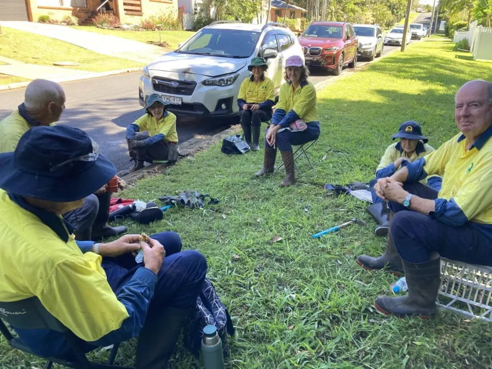 Landcare volunteers in hi-vis resting on camp chairs and grass beside parked cars during a break