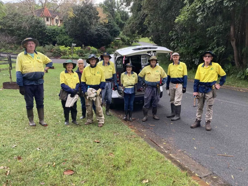 Group of Landcare volunteers in hi-vis shirts and boots standing beside a car on a quiet suburban street